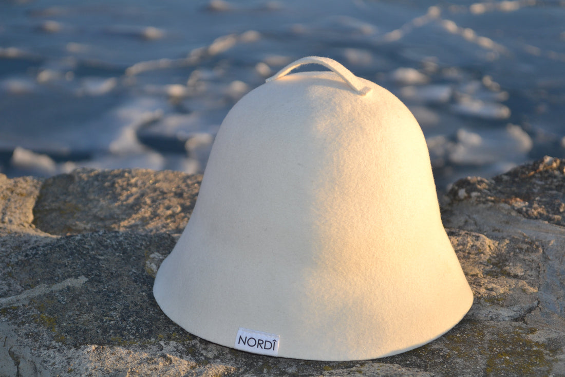 A signature NORDI wool sauna hat resting on a stone ledge by a cold lake, representing the traditional hot and cold therapy wellness ritual.